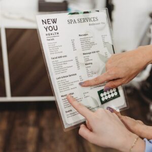 Hands pointing to a laminated spa services menu at New You Health, listing facials, massage, and lash and brow services available in Peoria.