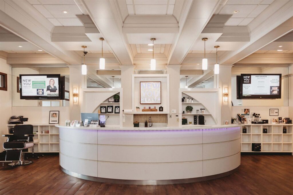Welcoming front desk at New You Health in Peoria, Illinois, featuring soft lighting, wood flooring, digital displays, and modern white decor with a glowing lavender accent.