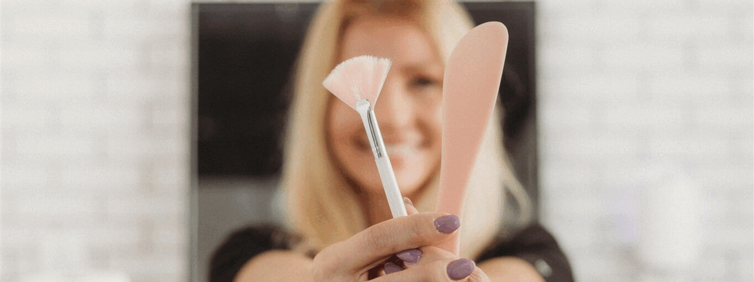 A smiling esthetician at New You Health holds up a pink facial brush and spatula toward the camera, showcasing skincare tools with a soft, blurred background.